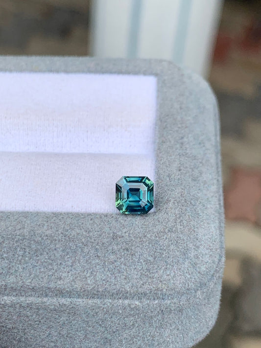Le Saphir Teal • 1,30ct de [NOUVEAU] Bonnot Paris, une pierre précieuse de taille carrée aux reflets bleus et verts, scintille sur une surface grise coussinée, ses facettes mises en valeur sur un fond flou.