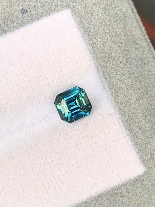 La [NOUVELLE] pierre précieuse Bonnot Paris Saphir Teal • 1,46 ct, de taille rectangulaire avec une teinte bleu-vert saisissante provenant des célèbres pierres précieuses de Madagascar, est présentée sur un fond texturé blanc et gris, mettant en valeur ses couleurs vibrantes et sa clarté à travers ses facettes réfléchissantes.