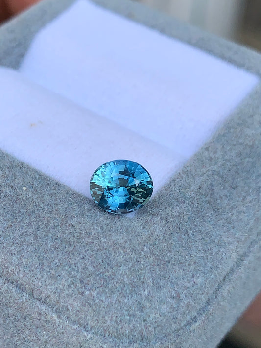 Le Saphir Teal de 1,56 carat de Bonnot Paris, originaire de Madagascar, scintille de ses reflets bleus sur un coussin gris, dans un écrin ouvert. Cette pierre précieuse rare est magnifiquement taillée en rond.