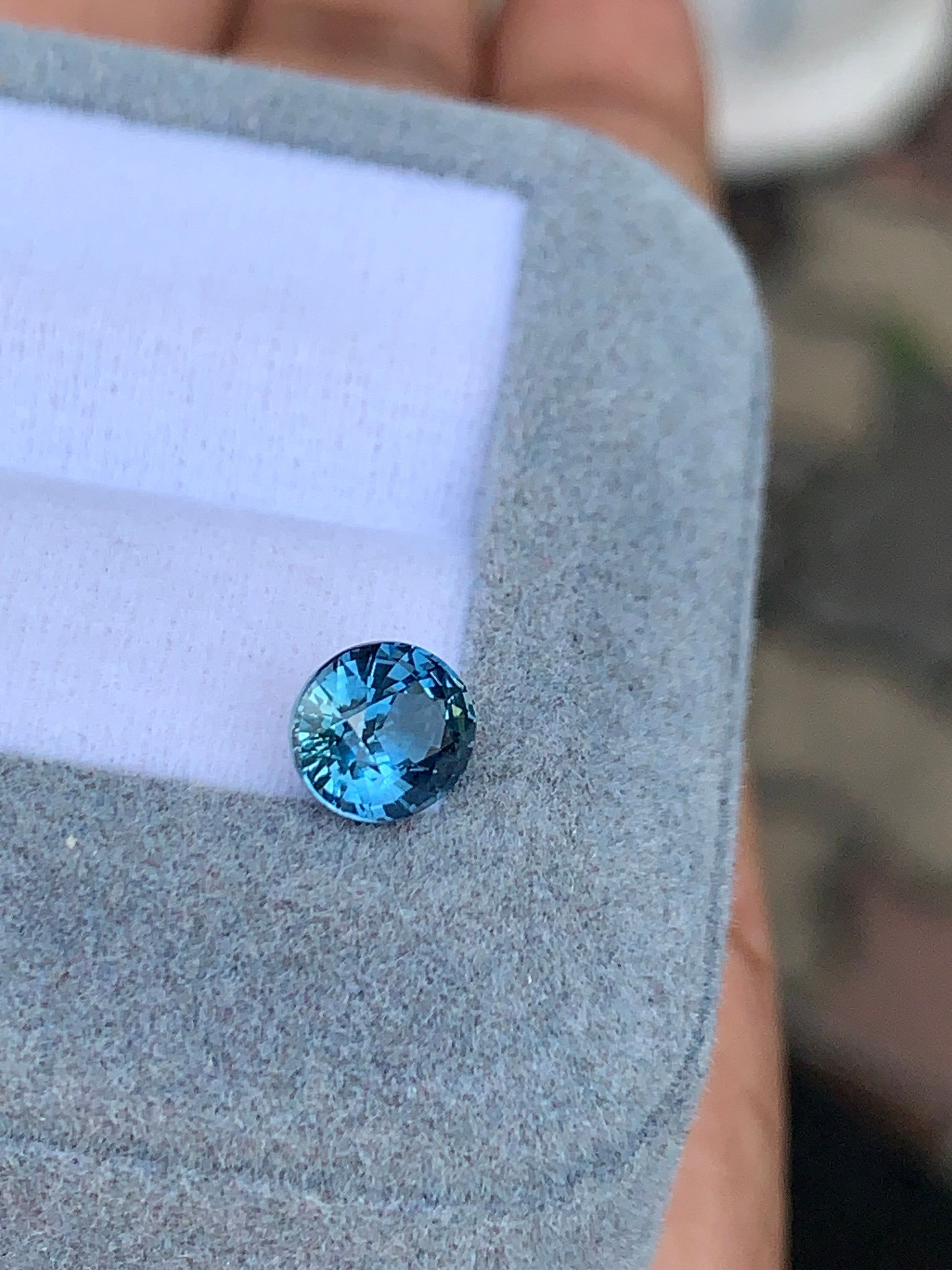 La pierre précieuse unique et ronde Saphir Teal de 1,56 ct de [NOUVEAU] Bonnot Paris, provenant de Madagascar, repose sur un coussin gris doux sur un fond blanc, rayonnant de nuances bleues vibrantes alors qu'elle est délicatement tenue dans la main de quelqu'un.