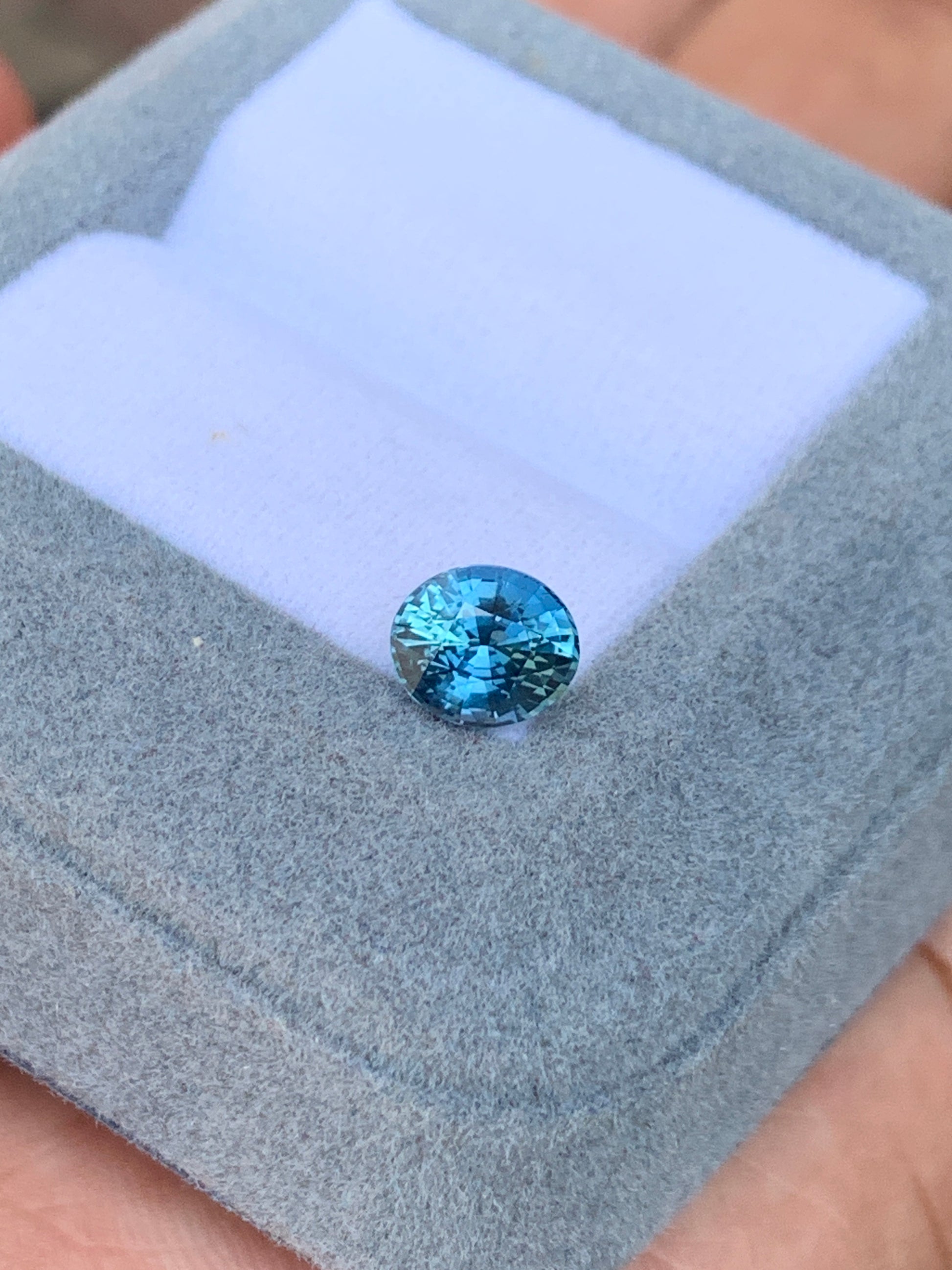 L'exquis Saphir Teal de 1,56 carat de Bonnot Paris, une pierre précieuse rare de Madagascar, est présenté dans un écrin gris ouvert doublé de tissu blanc. Cette pierre bleue ronde scintille de mille feux, mettant en valeur sa couleur vive et ses facettes saisissantes.
