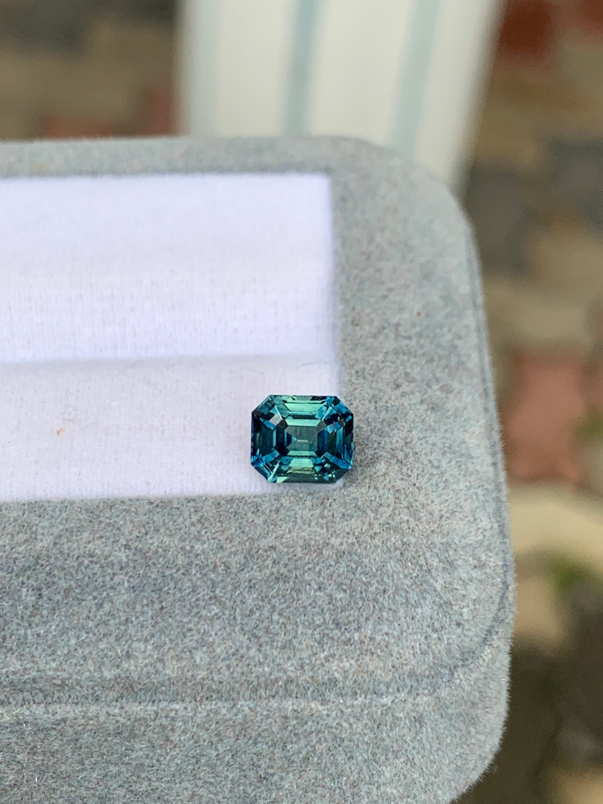 Le Saphir Teal de 1,57 carat de Bonnot Paris, une pierre précieuse naturelle de taille carrée, repose sur un tissu gris. Sa clarté éclatante et ses facettes réfléchissant la lumière rehaussent sa couleur vibrante sur un fond subtilement flou – une véritable gemme d'exception.