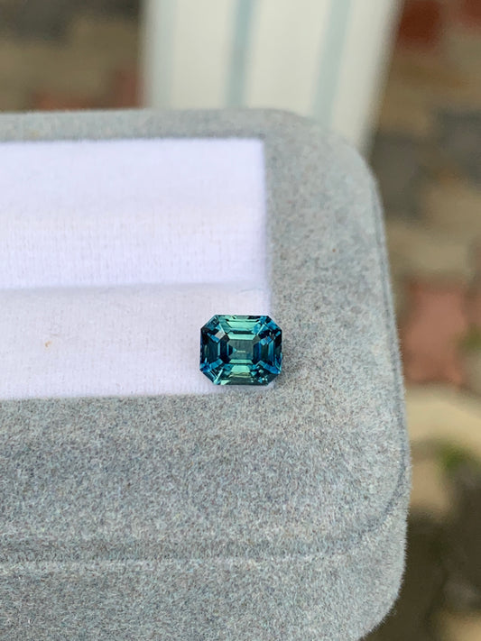 Le Saphir Teal de 1,57 carat de Bonnot Paris, une pierre précieuse naturelle de taille carrée, repose sur un tissu gris. Sa clarté éclatante et ses facettes réfléchissant la lumière rehaussent sa couleur vibrante sur un fond subtilement flou – une véritable gemme d'exception.
