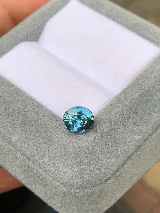 Une pierre précieuse Saphir Teal taille brillant • 1,57 ct de Madagascar de Bonnot Paris repose sur une boîte à bijoux gris clair et veloutée avec une doublure en tissu blanc, scintillant gracieusement à la lumière comme une pierre précieuse non chauffée.