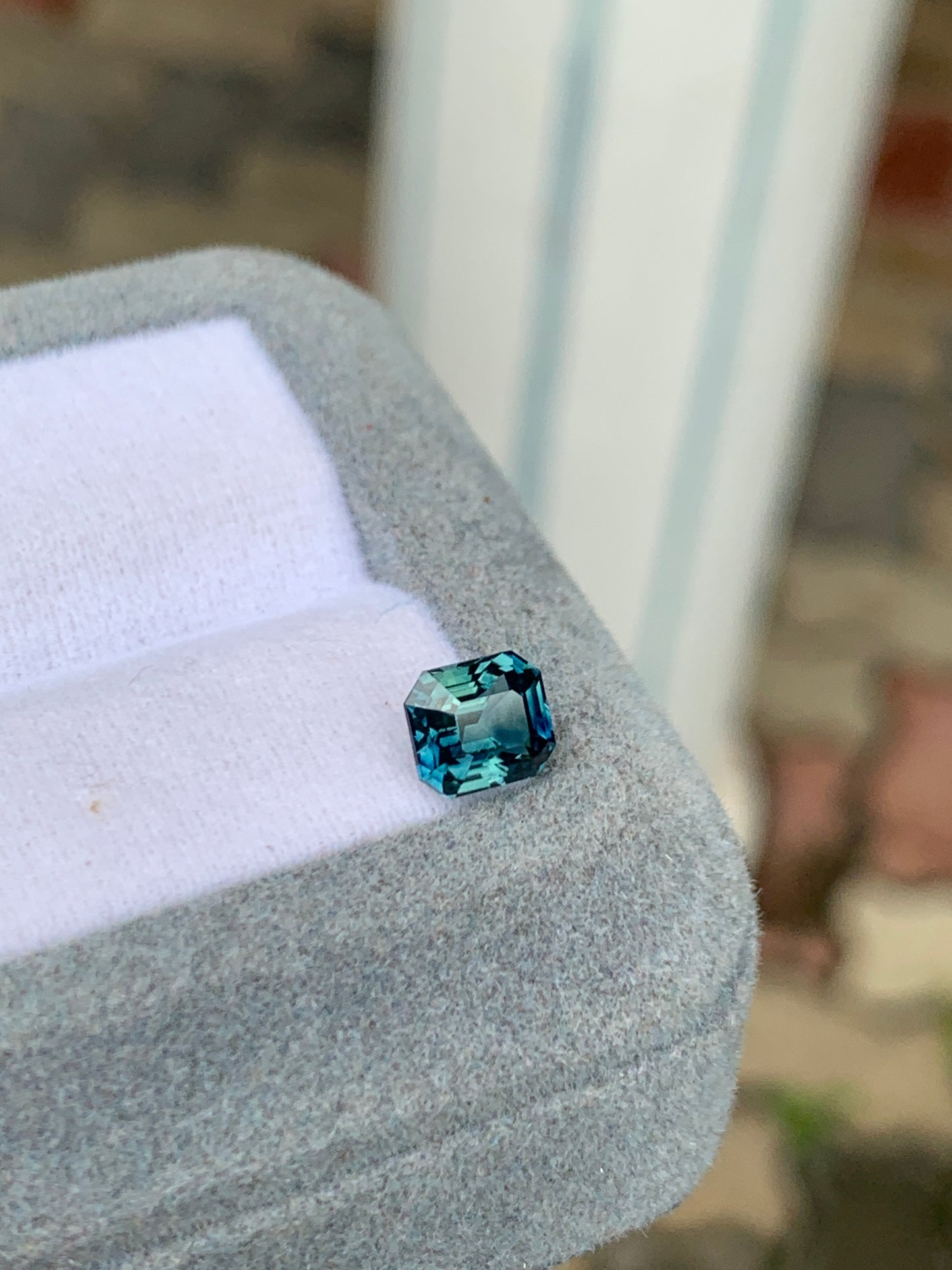 Le Saphir Teal • 1,57 carat de Bonnot Paris, une pierre précieuse bleue taille émeraude, est élégamment présenté dans un écrin de velours gris. Le fond flou vert et marron évoque un environnement naturel extérieur, soulignant l'élégance de la pierre.