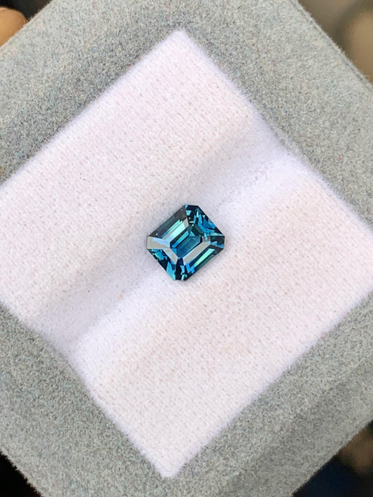 Le Saphir Teal • 1,58 ct de Bonnot Paris, une pierre précieuse bleue rectangulaire taille émeraude, est élégamment présenté sur un coussin blanc doux à l'intérieur d'un cadre gris, mettant en valeur une création joaillière raffinée.