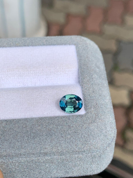 La pierre précieuse bleu sarcelle de Madagascar, Saphir Teal • 1,60 ct de Bonnot Paris, est présentée dans une boîte grise ouverte avec une doublure blanche sur un motif flou de briques imbriquées aux tons terre en arrière-plan.