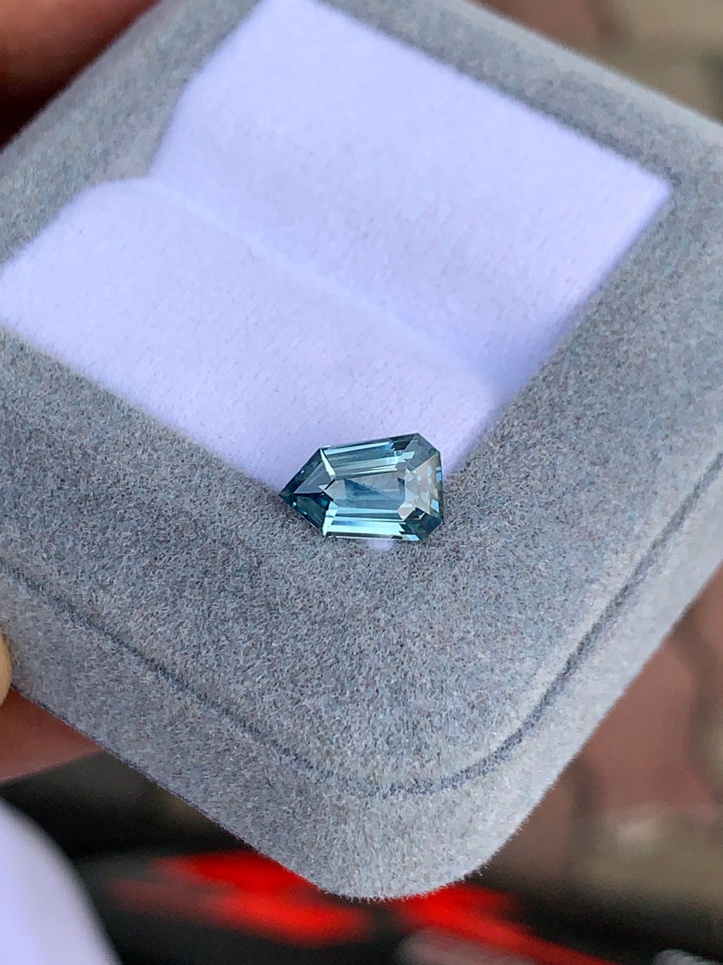 Dans un écrin ouvert recouvert d'un tissu gris clair moelleux se trouve le Saphir Teal, une pierre précieuse taille émeraude de 1,65 carat signée [NOUVEAU] Bonnot Paris. Sa teinte bleue scintillante rivalise avec un véritable bleu sarcelle, avec un fond flou qui accentue sa beauté naturelle et la précision de ses facettes.
