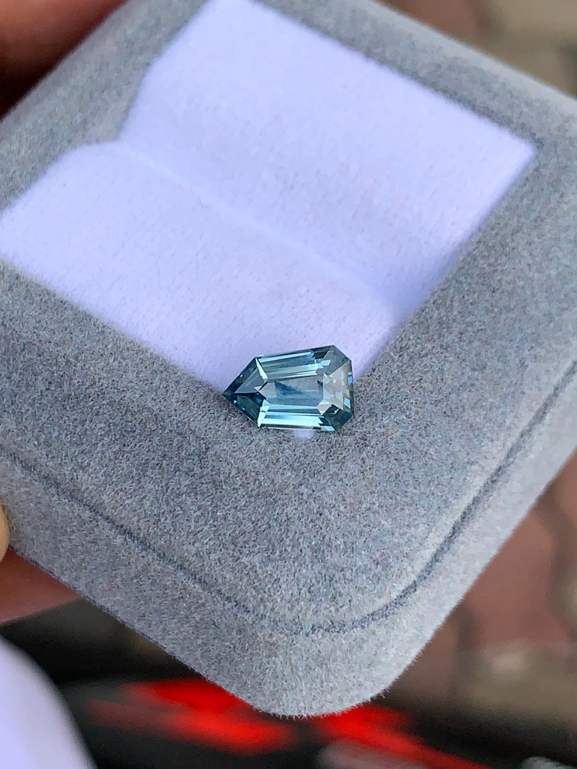 Dans un écrin ouvert recouvert d'un tissu gris clair moelleux se trouve le Saphir Teal, une pierre précieuse taille émeraude de 1,65 carat signée [NOUVEAU] Bonnot Paris. Sa teinte bleue scintillante rivalise avec un véritable bleu sarcelle, avec un fond flou qui accentue sa beauté naturelle et la précision de ses facettes.
