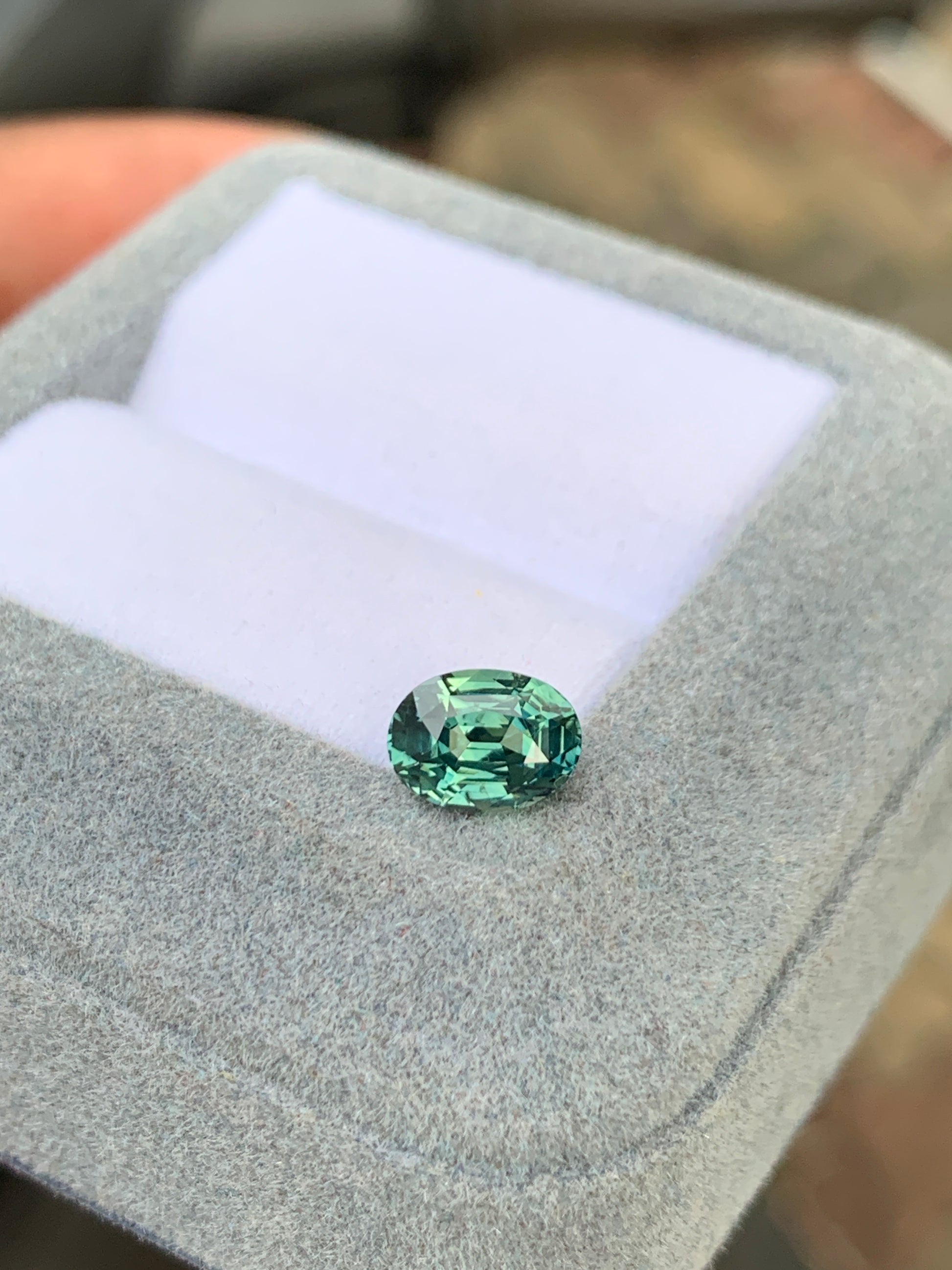 Un Saphir Teal de 1,65 carat de la marque Bonnot Paris [NOUVEAU] est présenté dans un écrin à bijoux gris clair matelassé. Le couvercle ouvert révèle un intérieur blanc où la pierre précieuse verte ovale non chauffée brille de ses facettes polies et de sa surface réfléchissante.
