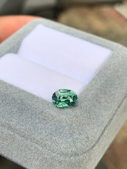 Un Saphir Teal de 1,65 carat de la marque Bonnot Paris [NOUVEAU] est présenté dans un écrin à bijoux gris clair matelassé. Le couvercle ouvert révèle un intérieur blanc où la pierre précieuse verte ovale non chauffée brille de ses facettes polies et de sa surface réfléchissante.