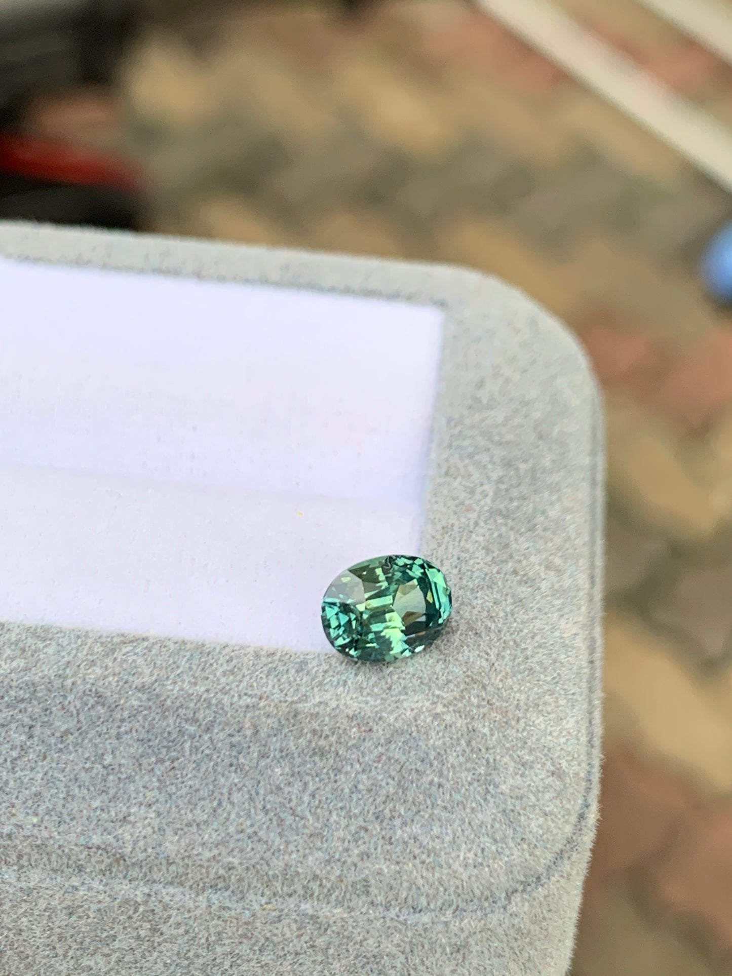 Le Saphir Teal de 1,65 ct de [NOUVEAU] Bonnot Paris, une exquise petite pierre précieuse ovale, repose élégamment sur une boîte recouverte de tissu gris doux, captant magnifiquement la lumière sur un fond multicolore flou.