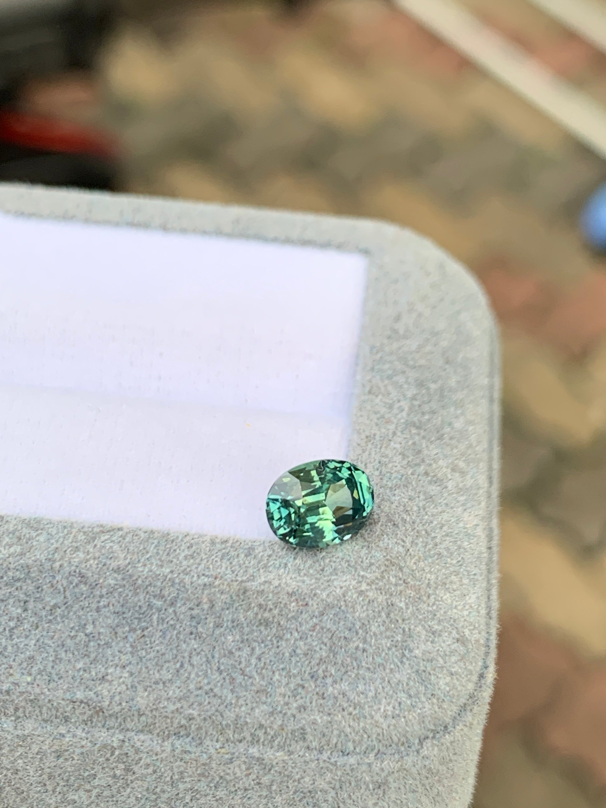 Le Saphir Teal de 1,65 ct de [NOUVEAU] Bonnot Paris, une exquise petite pierre précieuse ovale, repose élégamment sur une boîte recouverte de tissu gris doux, captant magnifiquement la lumière sur un fond multicolore flou.