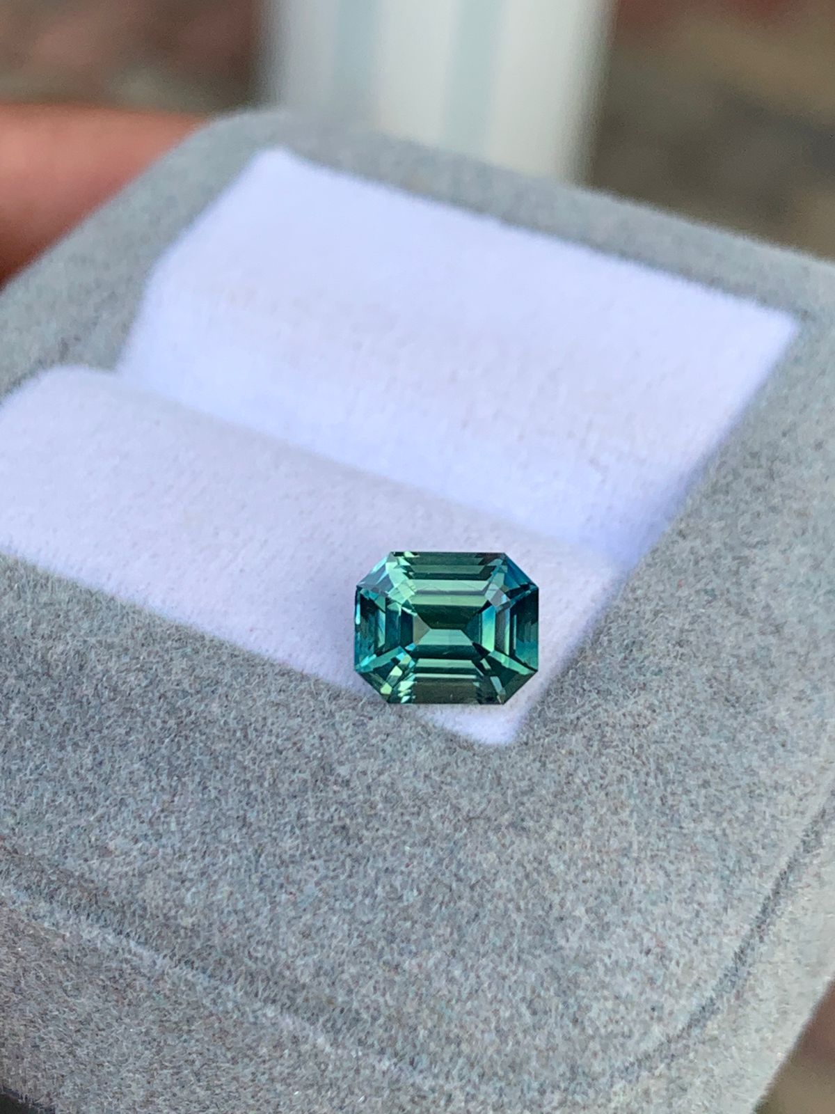 Les doigts d’une personne arborent le Saphir Teal • 2,01ct de [NOUVEAU] Bonnot Paris : une pierre précieuse verte taille émeraude, non chauffée et réfléchissant la lumière de ses facettes précises, sertie sur un écrin à bijoux gris avec une doublure blanche.