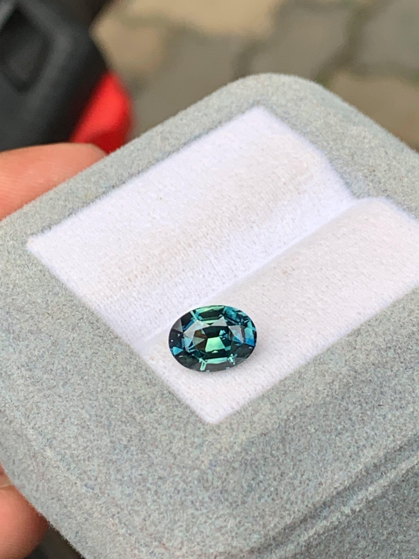 La pierre précieuse Saphir Teal 2,03 ct, de [NOUVEAU] Bonnot Paris, présente des teintes bleu et vert profonds, élégamment serties dans un coffret à bijoux gris avec une doublure blanche immaculée.