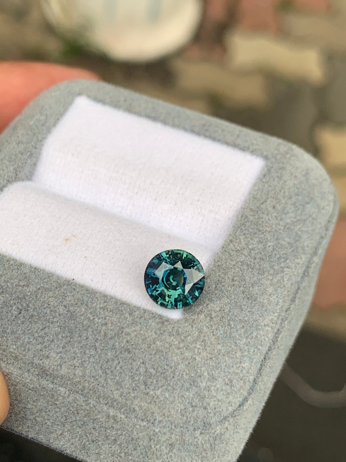 Un saphir bleu sarcelle de 2,53 carats de Bonnot Paris, évoquant les collections de haute joaillerie, est présenté dans un écrin gris ouvert doublé de tissu blanc. La pierre ronde bleu sarcelle scintille à la lumière naturelle, mettant en valeur ses facettes sur un fond flou.