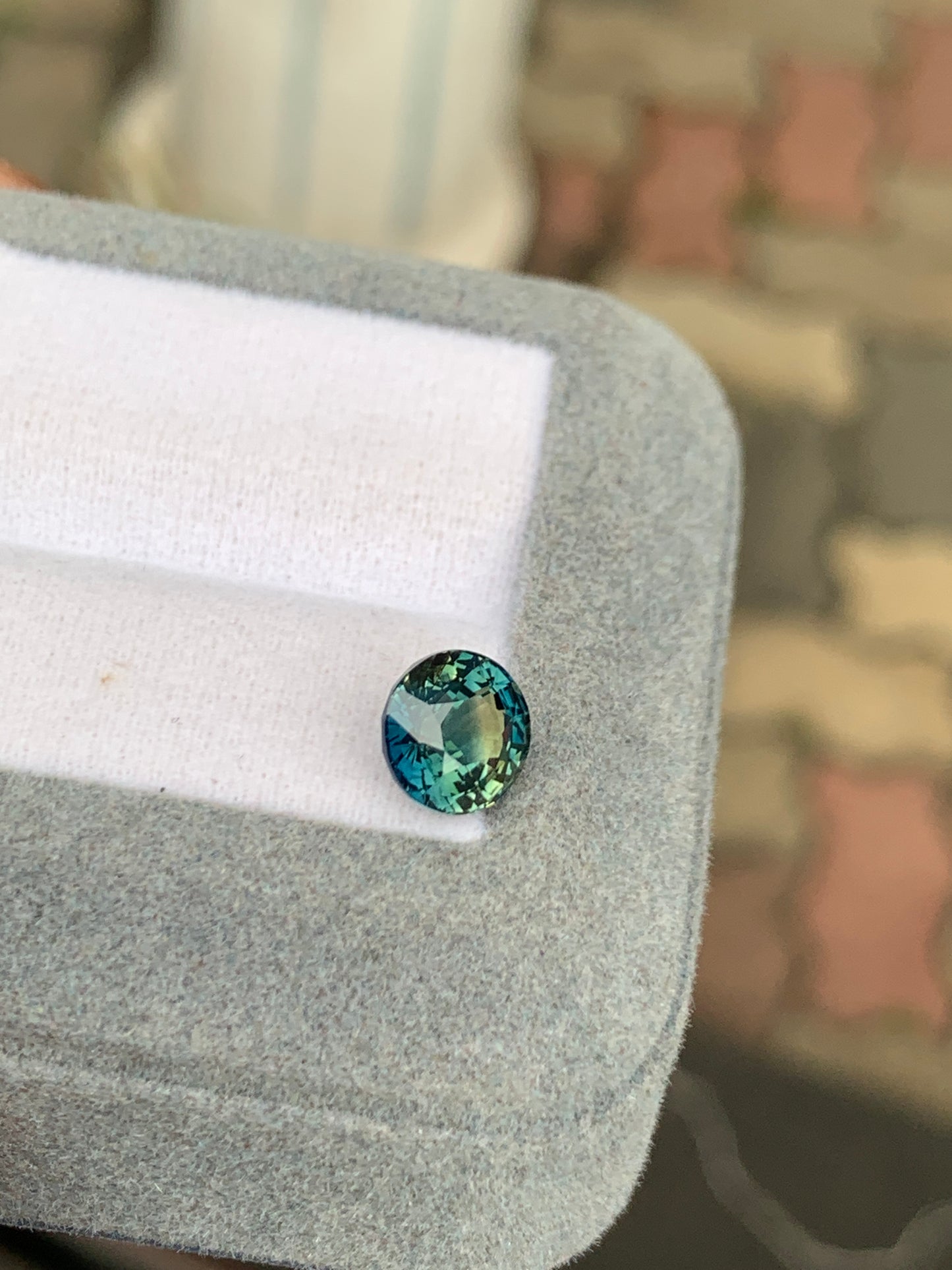 Le Saphir Teal • 2,53 ct de Bonnot Paris est une pierre précieuse ronde et facettée aux ravissantes nuances bleu sarcelle et vertes. Il est magnifiquement présenté dans un écrin gris doublé de blanc, sur un fond subtilement flouté qui souligne son élégance de haute joaillerie dans des tons doux.