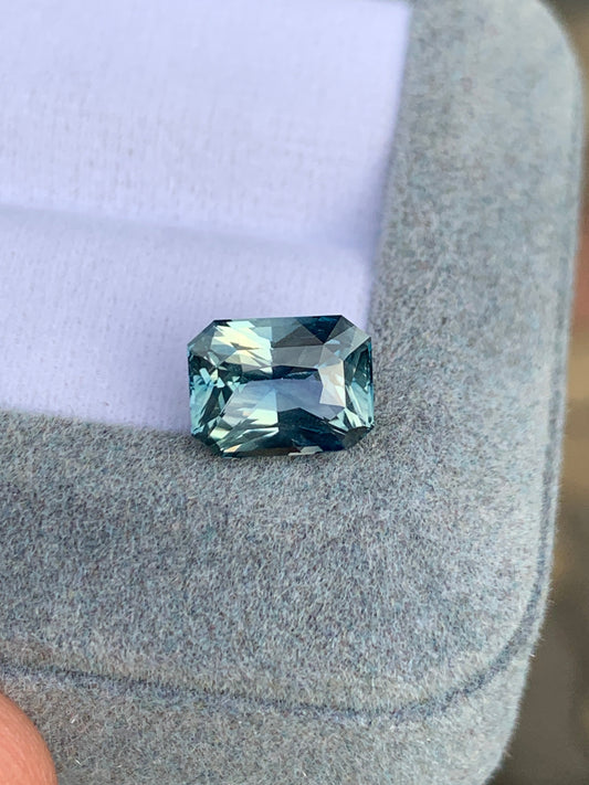 Une pierre précieuse Saphir Teal de 3,20 carats de Bonnot Paris, originaire de Madagascar, repose sur une surface gris clair et douce. Ses facettes rectangulaires scintillent, captant la lumière pour révéler de magnifiques teintes variées.