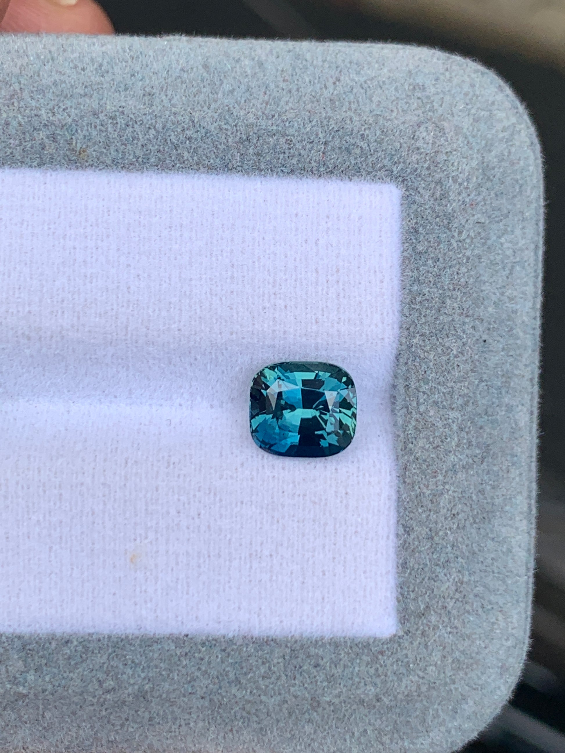 Un superbe Saphir Teal de 2,50 carats de Bonnot Paris de Madagascar, avec une taille coussin rectangulaire, repose sur un fond blanc à l'intérieur d'une boîte doublée de tissu gris, ses facettes captant magnifiquement la lumière et révélant des nuances bleues envoûtantes.