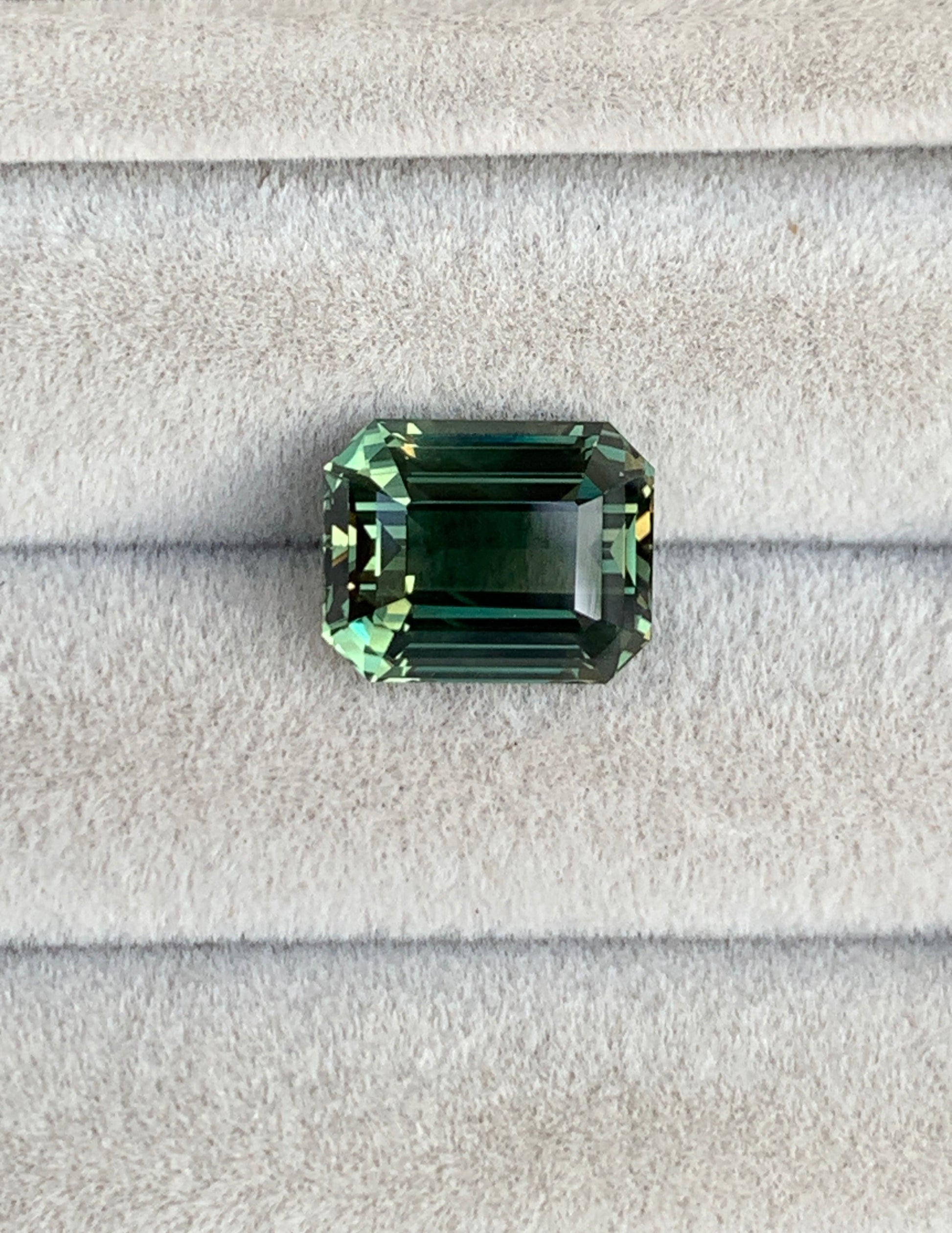 Le Saphir Vert • 6,30ct de Bonnot Paris est une pierre précieuse verte taille émeraude aux facettes complexes et à la teinte profonde et vibrante, élégamment présentée sur du velours gris.