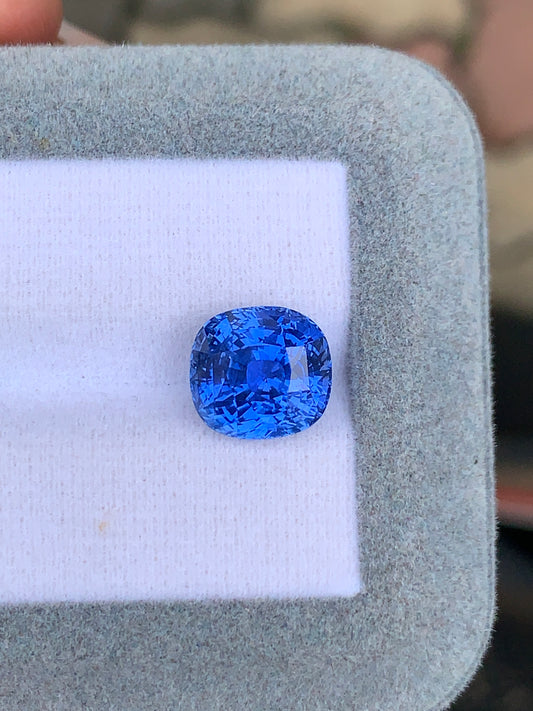 Un Saphir bleu de 7,08 carats de [NOUVEAU] Bonnot Paris est présenté sur un tissu gris clair, affichant en évidence sa couleur bleu vif et ses facettes complexes.