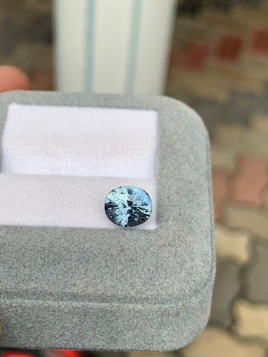 Un saphir gris bleu rond de 3,10 carats de Madagascar de Bonnot Paris scintille sur une base coussinée blanche à l'intérieur d'un coffret en velours gris, avec des pavés terreux flous en arrière-plan pour souligner son élégance.