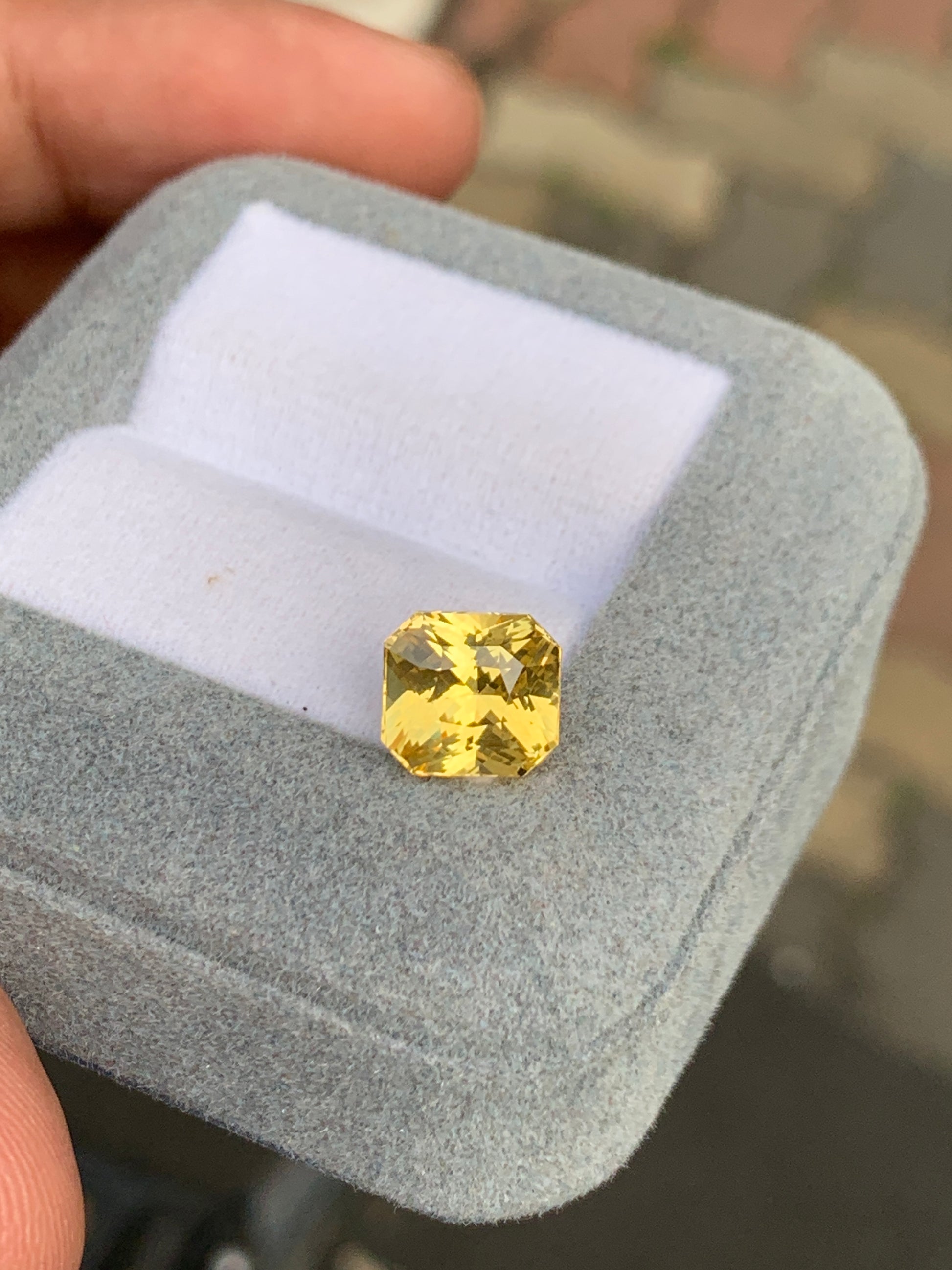 Une pierre précieuse taille coussin Saphir jaune Bonnot Paris de 4,00 ct est élégamment présentée dans un coffret à bijoux en velours gris avec un intérieur blanc, délicatement tenu dans une main sur fond de scène de trottoir sri-lankais légèrement floue.