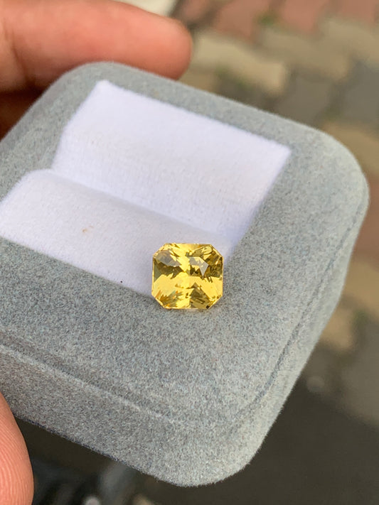 Une pierre précieuse taille coussin Saphir jaune Bonnot Paris de 4,00 ct est élégamment présentée dans un coffret à bijoux en velours gris avec un intérieur blanc, délicatement tenu dans une main sur fond de scène de trottoir sri-lankais légèrement floue.