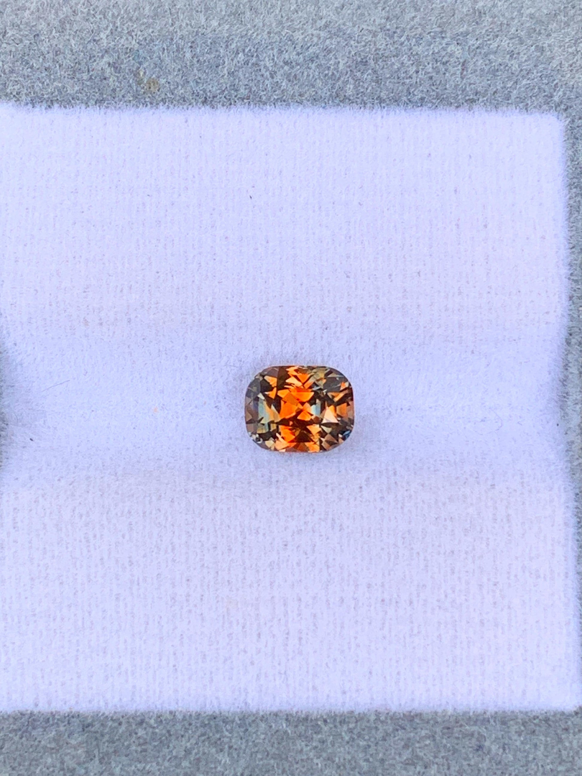 Une pierre précieuse orange taille coussin de 1,12 carat de la collection Saphir Multicolore de Bonnot Paris scintille sur une surface blanche coussinée, évoquant le monde vibrant des pierres précieuses du Sri Lanka. Elle reflète magnifiquement la lumière, dévoilant une palette de teintes orange et jaunes sur un fond gris.