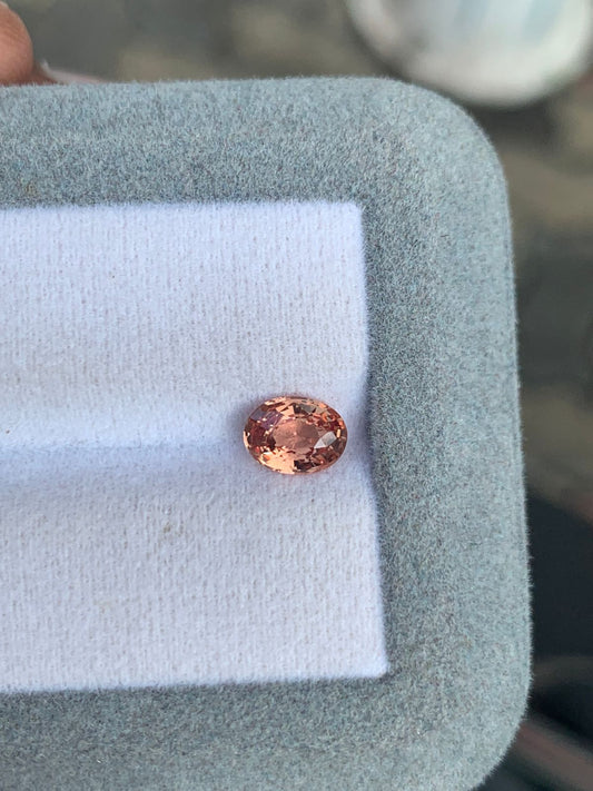Le [NOUVEAU] Saphir Padparadscha Bonnot Paris • 1,05 ct est une petite gemme ovale de Madagascar. Sa teinte pêche réfléchissante est mise en valeur sur un tissu blanc dans un cadre gris clair, positionné au centre de l'image.