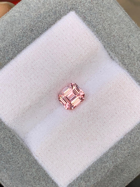 Le Saphir pêche • 1,40ct de Bonnot Paris, une pierre précieuse rose taille carrée, est élégamment présenté sur un coussin blanc dans un écrin gris, mettant en valeur ses facettes et son éclat.