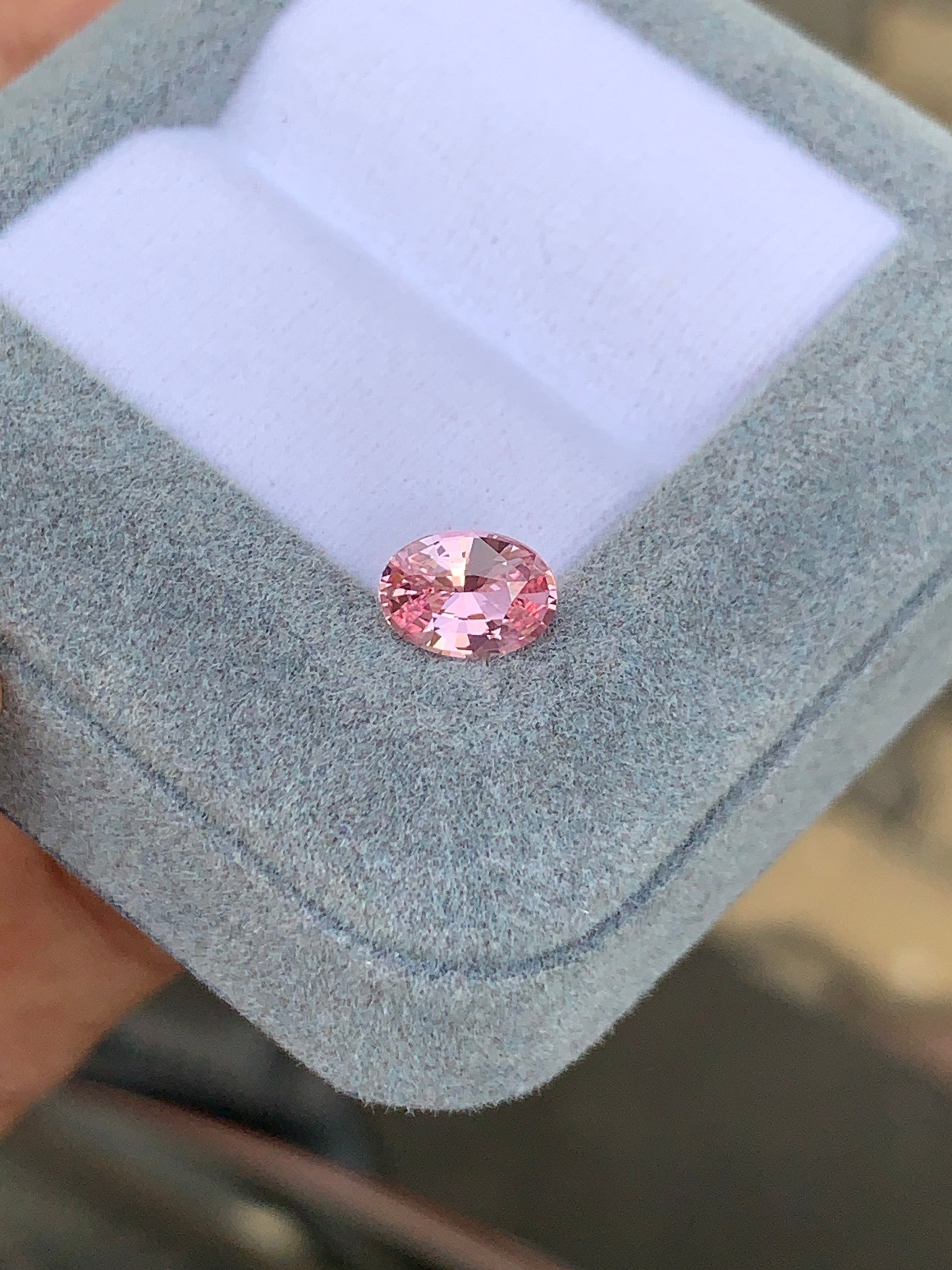 Le Saphir rose • 1,2ct de Bonnot Paris, une pierre précieuse rose de forme ovale à l'éclat brillant, est présenté dans un écrin gris ouvert à l'intérieur blanc, tenu à la main sur un fond flou.