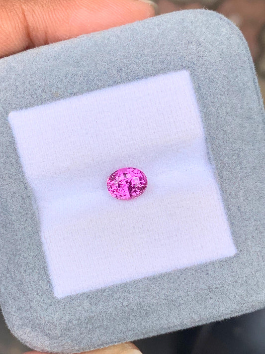 Une main tient un coffret à bijoux gris de [NOUVEAU] Bonnot Paris contenant un saphir rose de 1,51 carat. Serti sur un coussin blanc, ce saphir rose rond capte la lumière grâce à ses facettes, créant un effet scintillant.