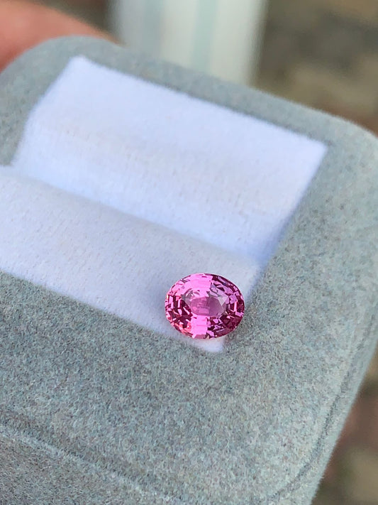 Un Saphir rose • 1,55ct de chez [NOUVEAU] Bonnot Paris repose radieusement sur un coussin blanc à l'intérieur d'un écrin à bijoux gris, sur un fond flou.