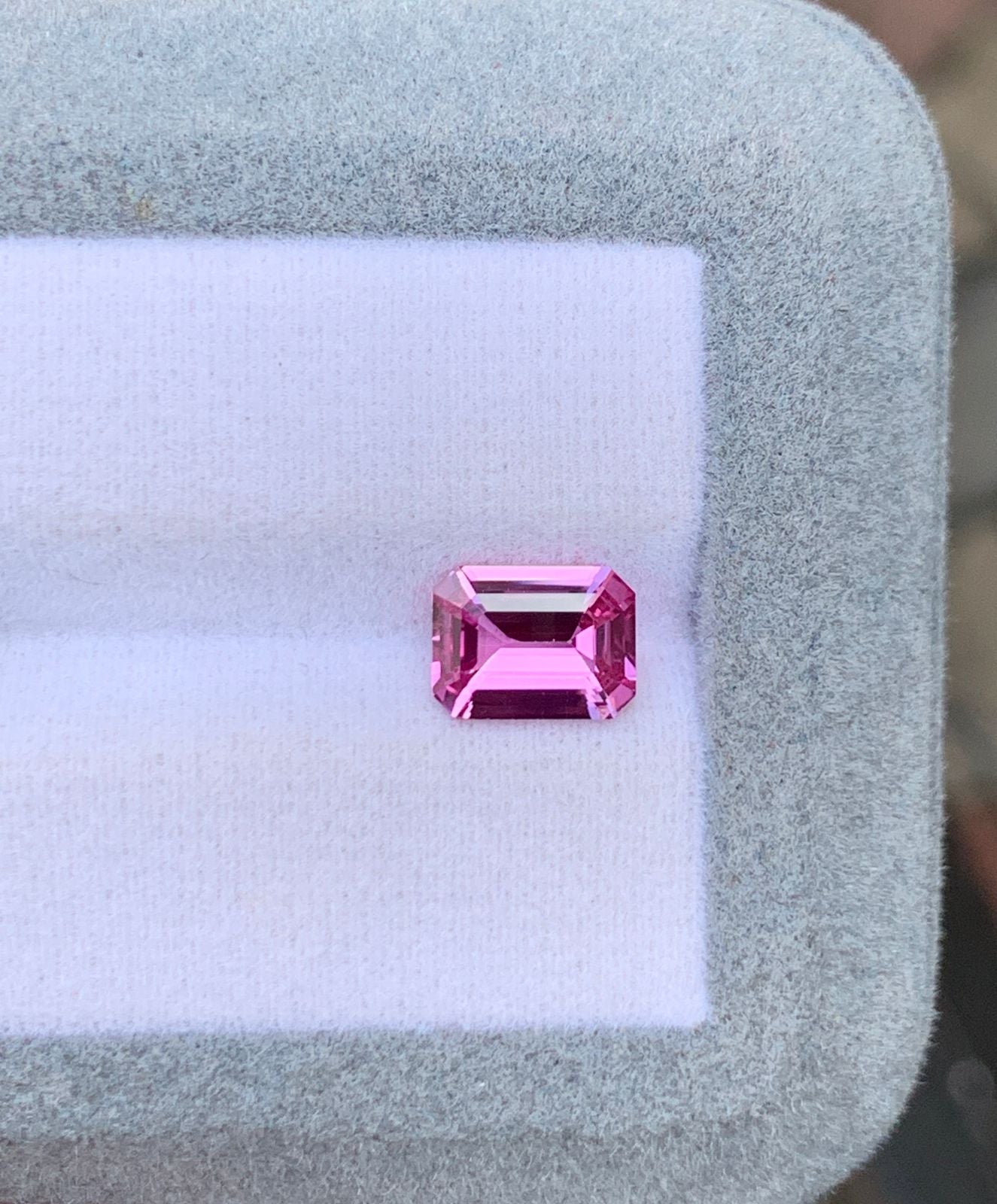 Un Saphir rose taille émeraude étincelant de 2,03 ct de Bonnot Paris est présenté sur un coussin blanc bordé de gris clair. Ses facettes captent la lumière, soulignant sa clarté et sa couleur vive, typiques des pierres précieuses.