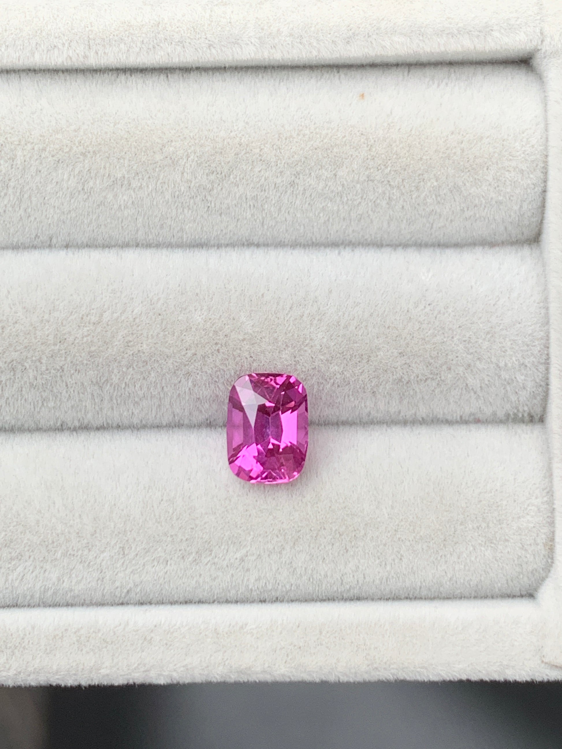 Un Saphir rose éclatant de 2,10 carats, taille coussin, signé Bonnot Paris, repose dans un écrin gris clair strié. Ses facettes captent la lumière, révélant de multiples nuances de rose.