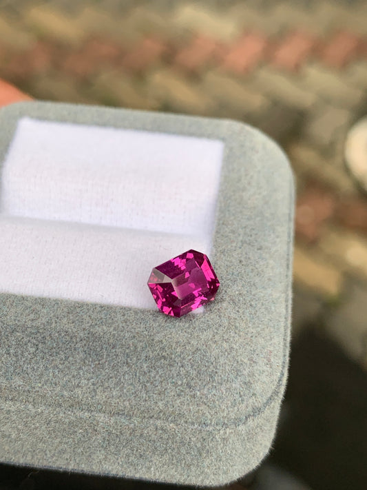 Un saphir rose vibrant de 3,02 ct avec une taille émeraude de Bonnot Paris est élégamment présenté sur une boîte à bijoux en velours gris clair, avec un fond flou aux tons terreux.