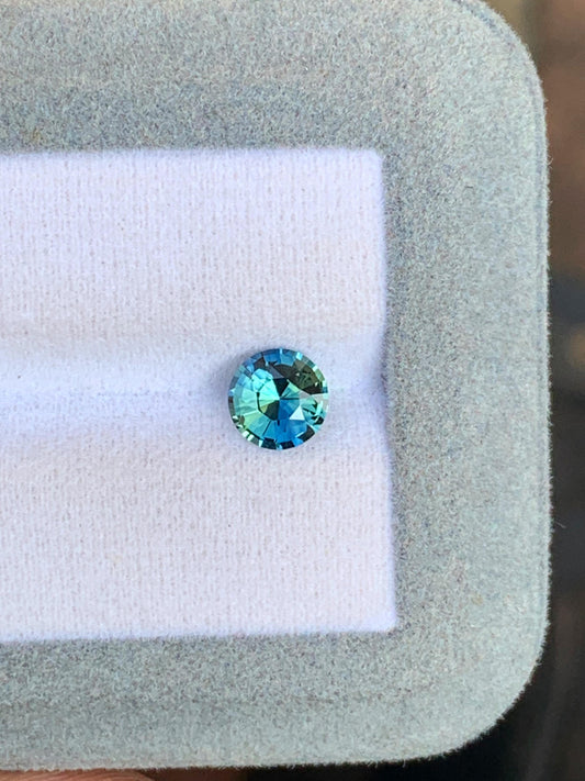 Le Saphir Teal (1,25 carat) de Bonnot Paris, une pierre ronde facettée, présente des nuances bleues et vertes envoûtantes sur une surface blanche et grise douce, mettant en valeur ses couleurs vibrantes. Cette pierre précieuse exquise captive par sa beauté naturelle rappelant les saphirs enchanteurs de Madagascar.