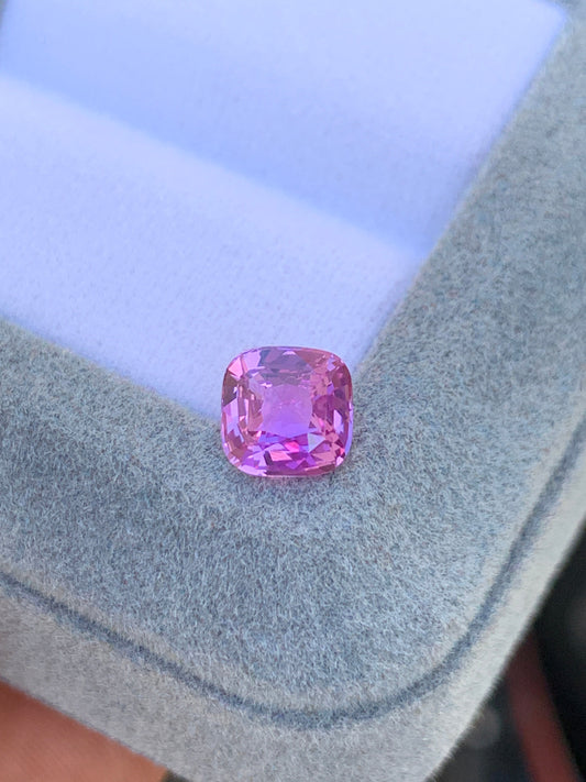Une pierre précieuse Saphir Rose taille coussin • 1,52 ct de Bonnot Paris brille de mille feux sur une surface grise douce, probablement une boîte à bijoux, avec ses facettes rehaussant sa teinte vibrante.
