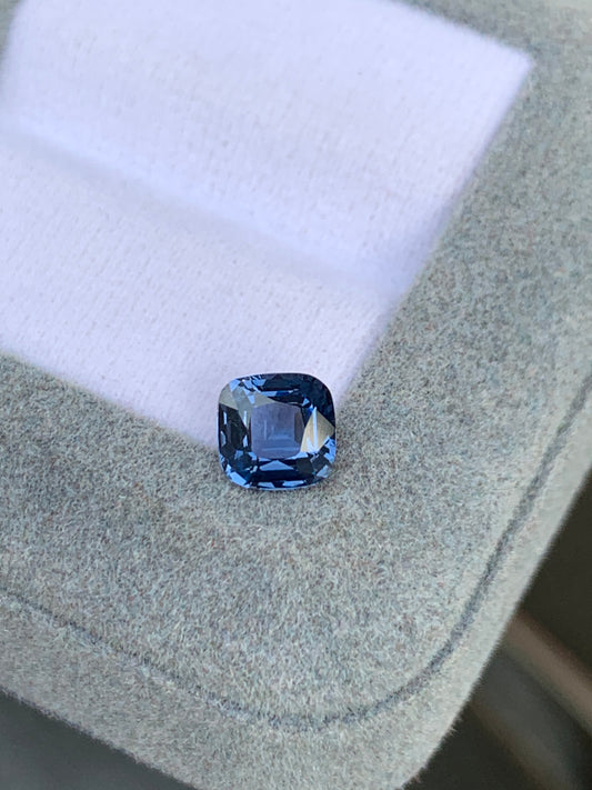 Le Spinelle bleu • 1,28 ct de [NOUVEAU] Bonnot Paris, une pierre précieuse taille coussin de Madagascar, repose sur un tissu gris clair sur un fond blanc doux. Sa surface facettée, d'une rare brillance, est réfléchissante.
